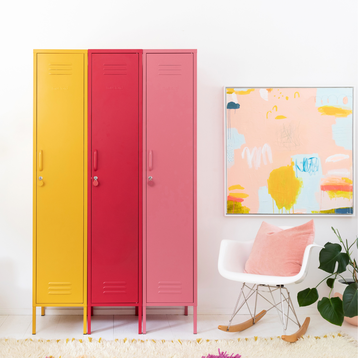 Three Mustard Made Skinny lockers stand side by side. Each is a different colour. From the left it is Mustard, Poppy and then Berry. To the right of the lockers are a large, framed artwork and a white rocking chair with a pink cushion on top. 