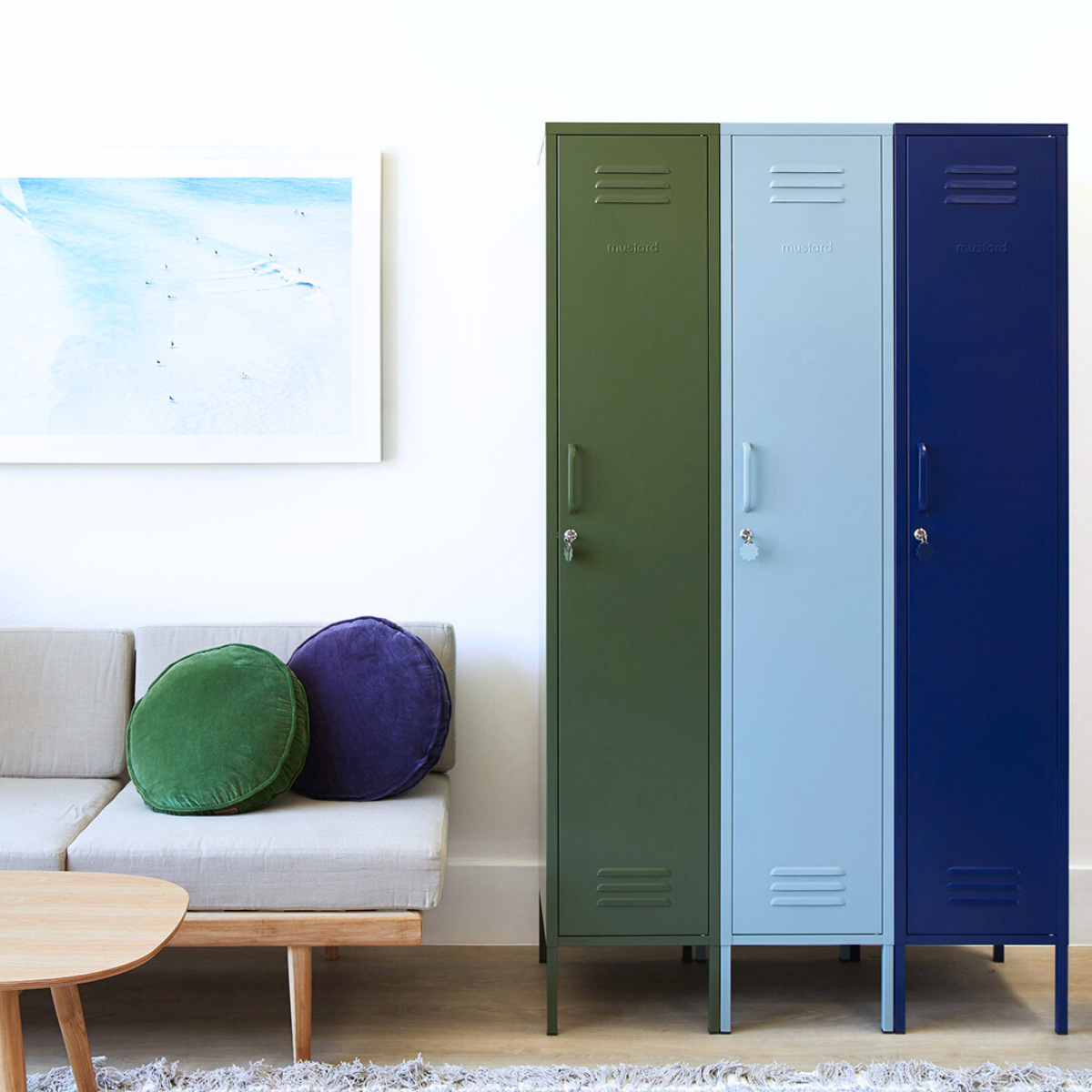 Three Mustard Made Skinny lockers stand side by side. Each is a different colour. From the left it is Olive, Ocean and then Navy. To the left of the lockers, a blue-toned artwork hangs on the wall. There is also a sofa with a timber frame, beige padding and two round cushions sitting on top — one blue and the other green. 