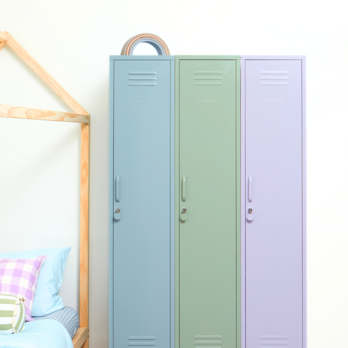 Three Mustard Made Skinny lockers stand side by side. Each is a different colour. From the left it is Ocean, Sage and then Lilac. To the left of the lockers is a bed with a wooden frame, and pastel bed linen. 