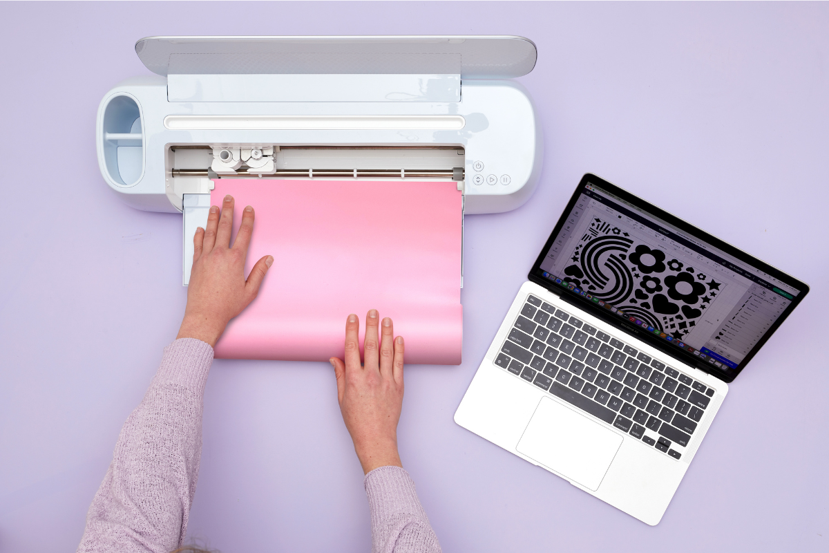 A close up of a person's hands feeding a sheet of pastel pink vinyl into a Cricut machine.