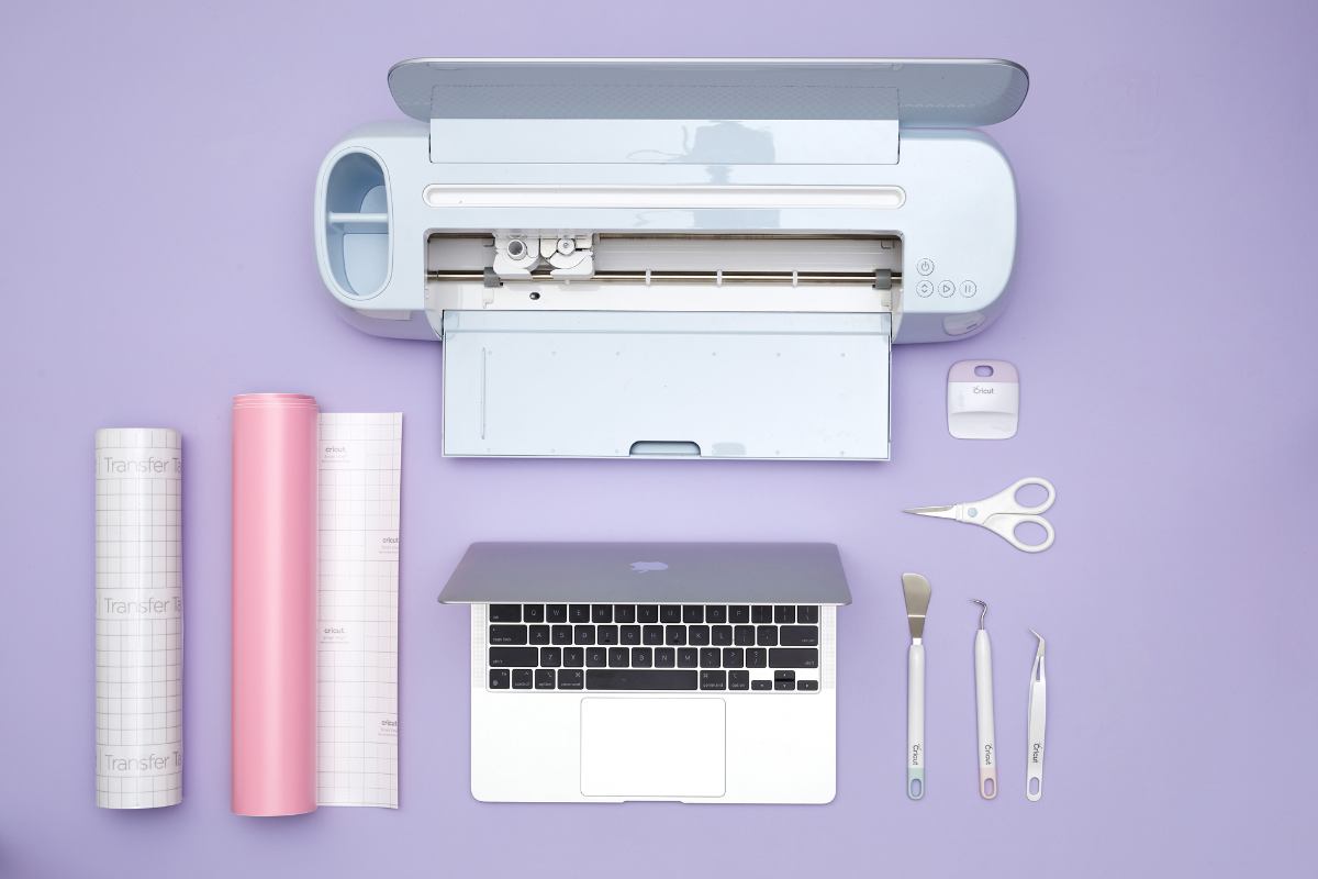 A flat lay with a lilac background shows a Cricut machine and laptop along with rolls of vinyl, scissors and Cricut tools.