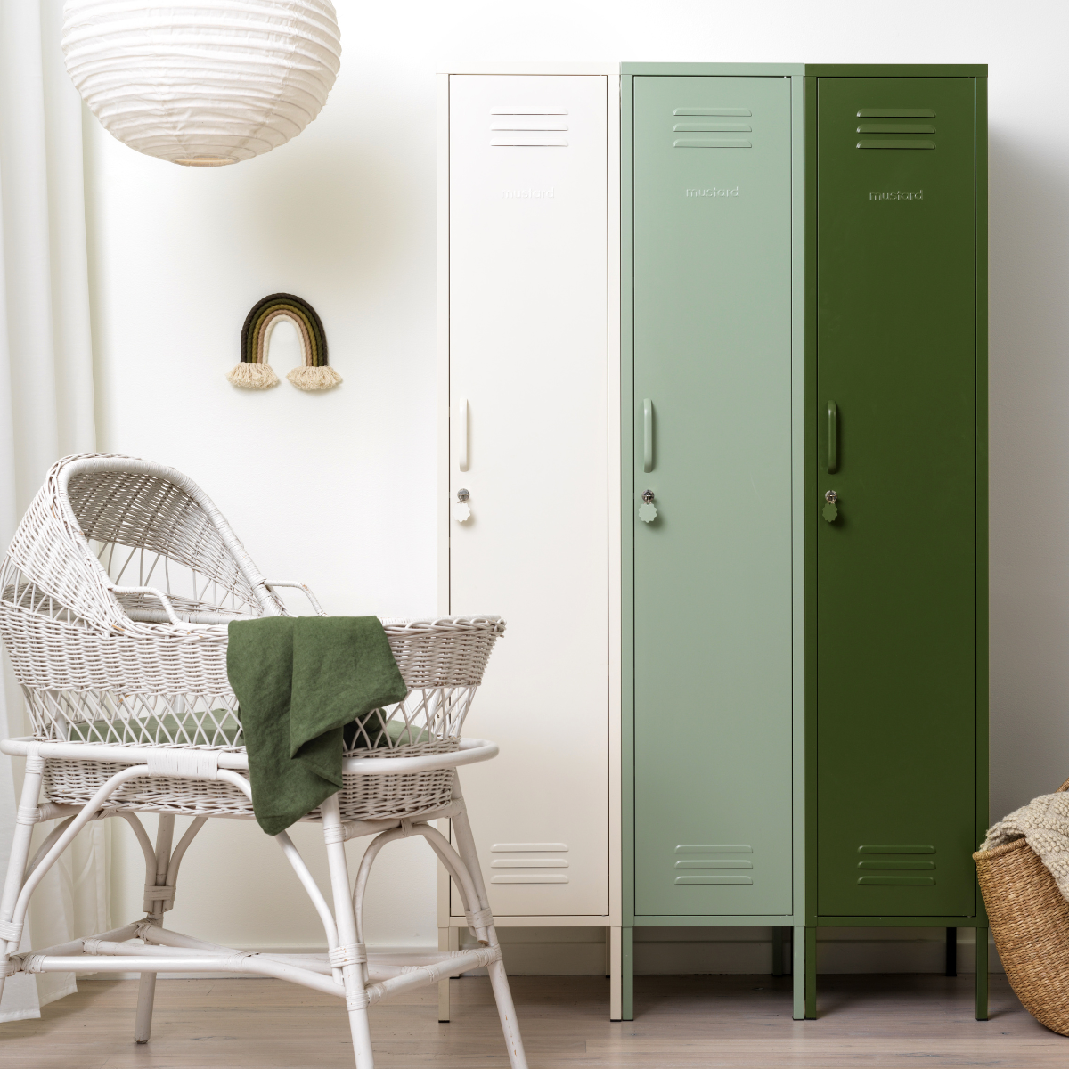 Three Mustard Made Skinny lockers stand side by side. Each is a different colour. From the left, it is White, Sage and then Olive. To the left of the lockers is a white, rattan bassinet with light green bed linen. A large round pendant light hangs above this. 