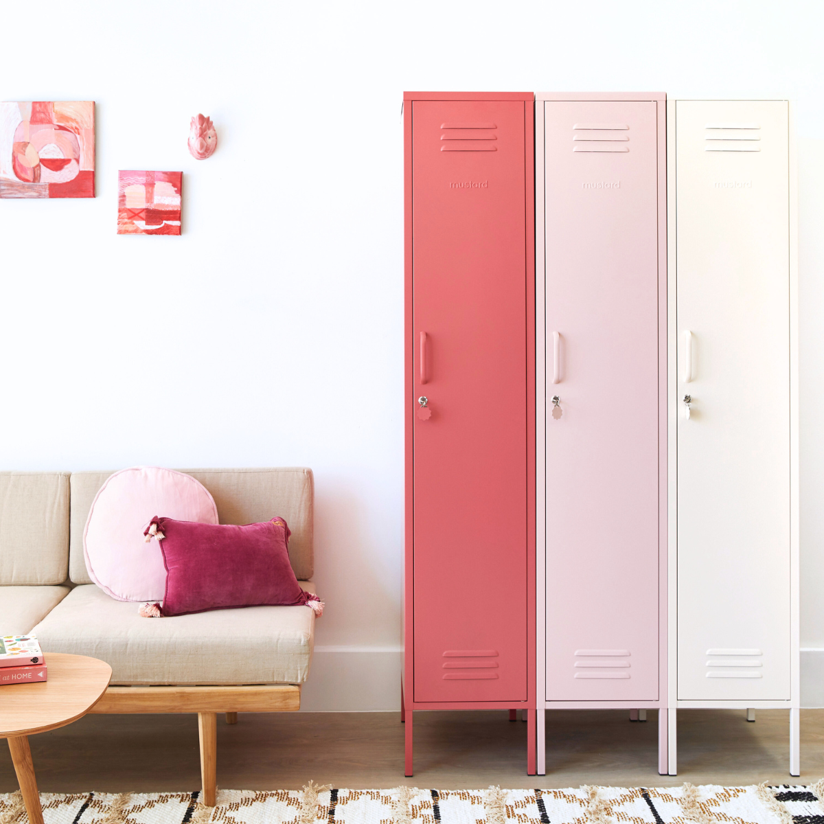 Three Mustard Made Skinny lockers stand side by side. Each is a different colour. From the left it is White, Blush and then Berry. To the left of the lockers, two pink-toned artworks hang on the wall. There is also a sofa with a timber frame, beige padding and two pink cushions sitting on top. 