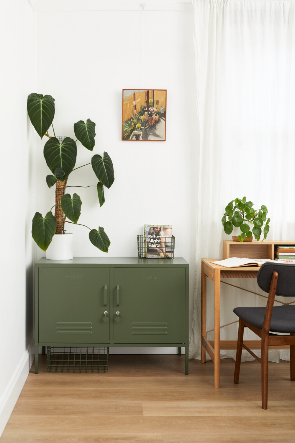 An Olive Lowdown is styled in a home office next to a small wooden desk. There is a tall potted plant on top of the locker and a small painting on the wall.