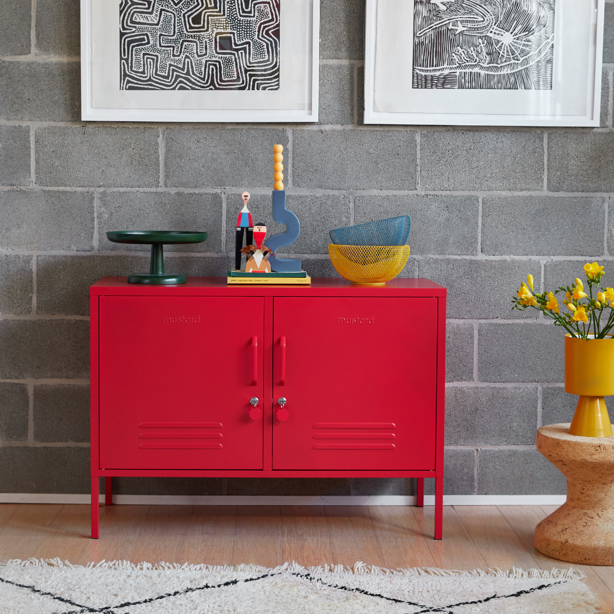 A Poppy Lowdown sits in front of a grey wall with monochrome art work hung on it. A vase of yellow flowers sits next to it and blue and yellow accessories decorate the top.