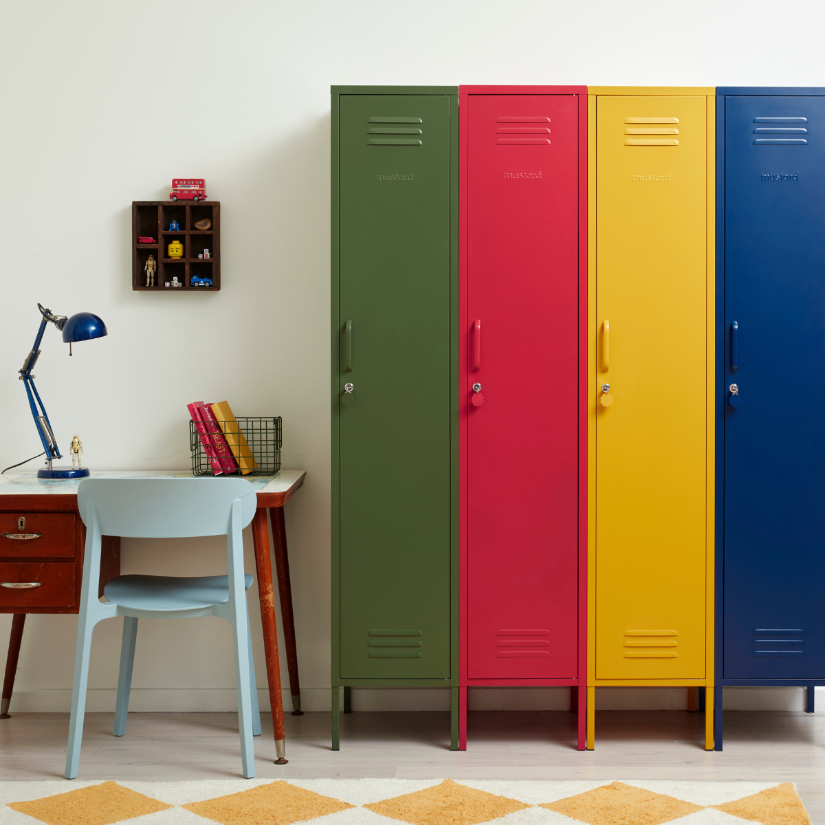 Four brightly coloured Skinny lockers sit side by side to create a rainbow of primary colours. Next to them is a small wooden desk with a pale blue chair.