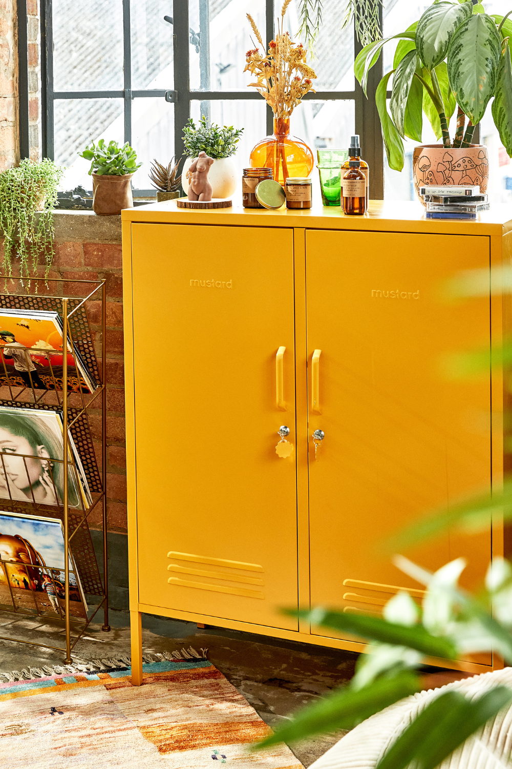 A Mustard Midi sits against an exposed brick wall below an industrial style window. There are books, plants and records scattered around.