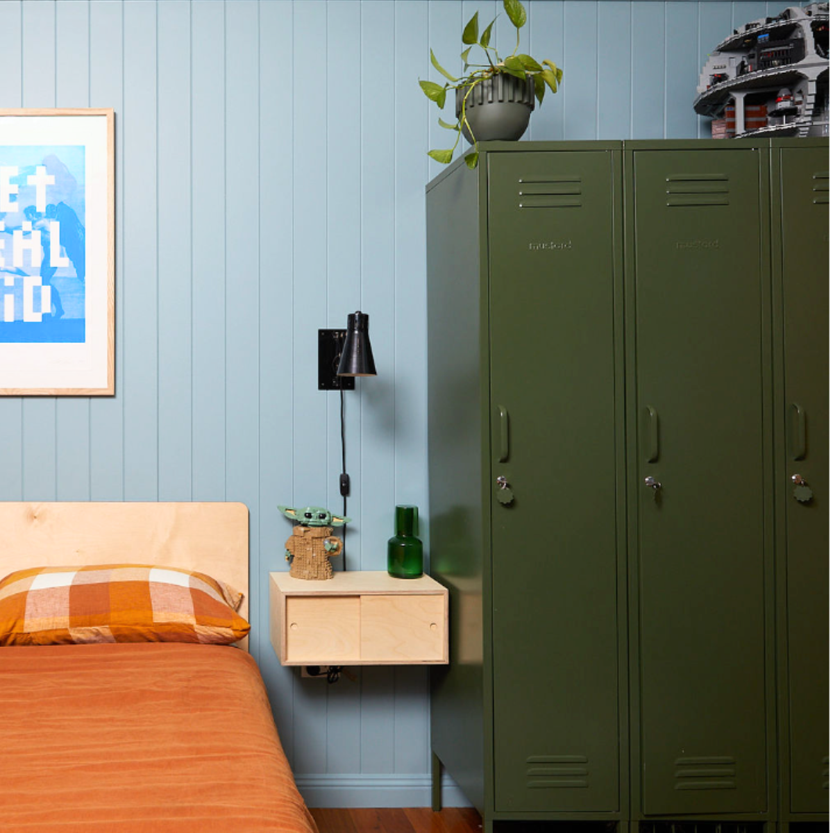 Three Mustard Made Skinny lockers stand side by side. They are all the colour, Olive. They are in front of a light blue-panelled wall. There is a single bed to the left of the lockers, with orange bed linen. A plywood floating bedside table sits beside the bed. There is a light fixture and Baby Yoda Lego figurine on top. 