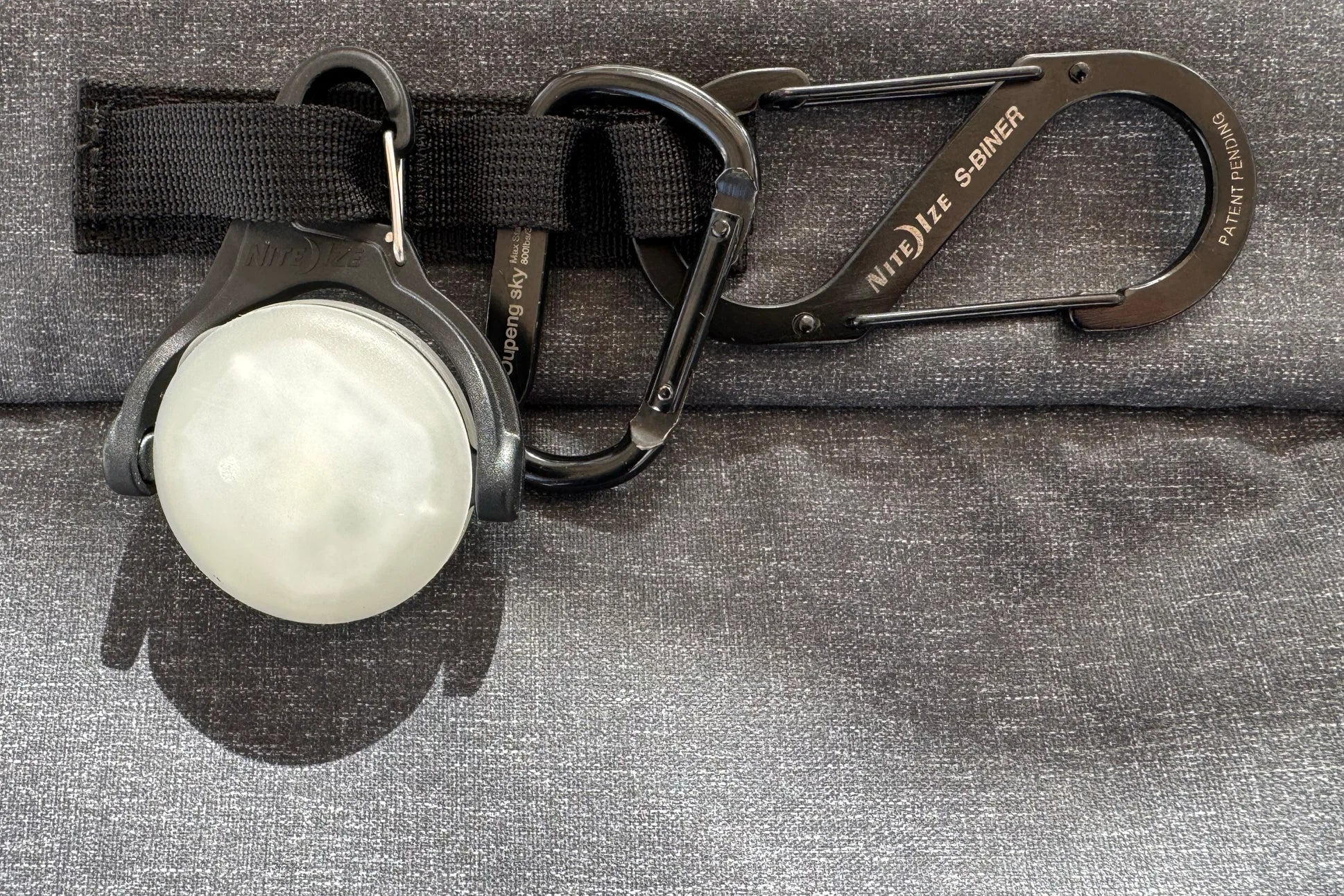 A Nite Ize S-Biner and light accessory attached to a black strap on a gray fabric background.