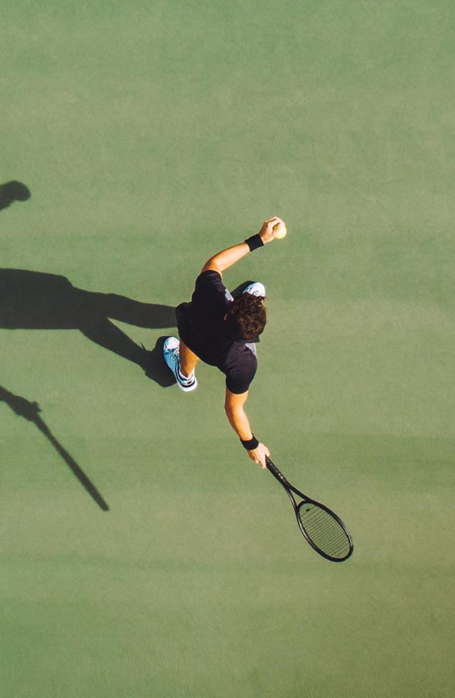 A tennis player holdin a black racket in his right hand and preparing to shot a ball that he holds in his other hand. A view from a bird perspective.