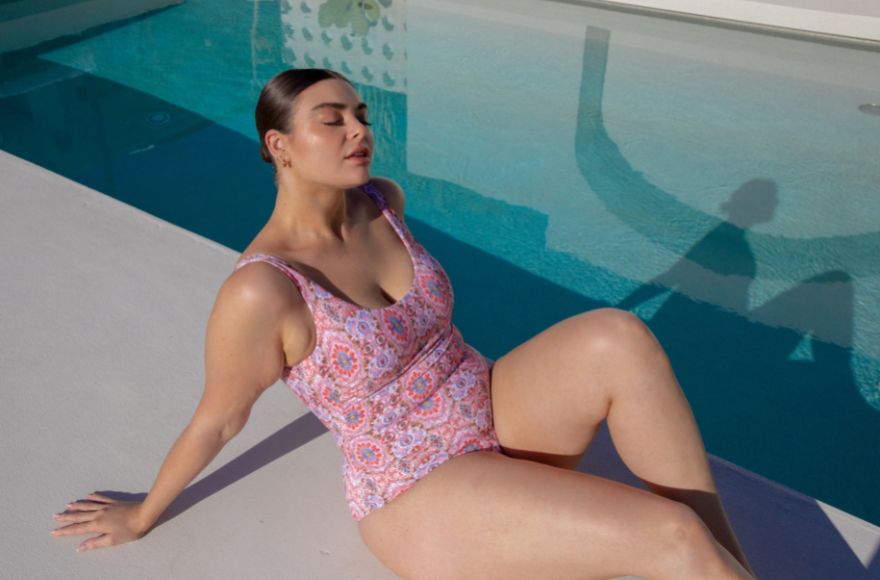 Brunette woman sits by the pool wearing Amalfi Pink boho print scoop one piece swimsuit