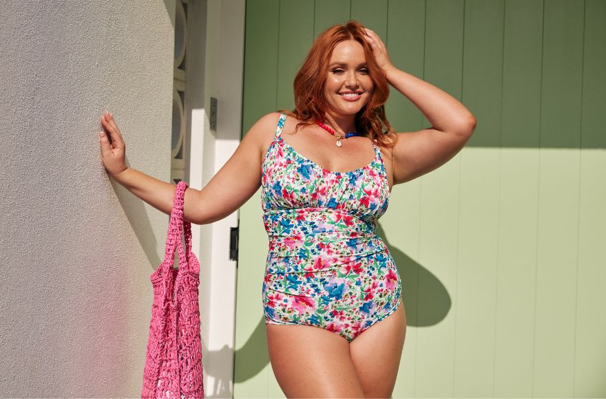 Woman with red hair poses in front of a pale green door wearing bright floral swimsuit holding a hot pink woven bag