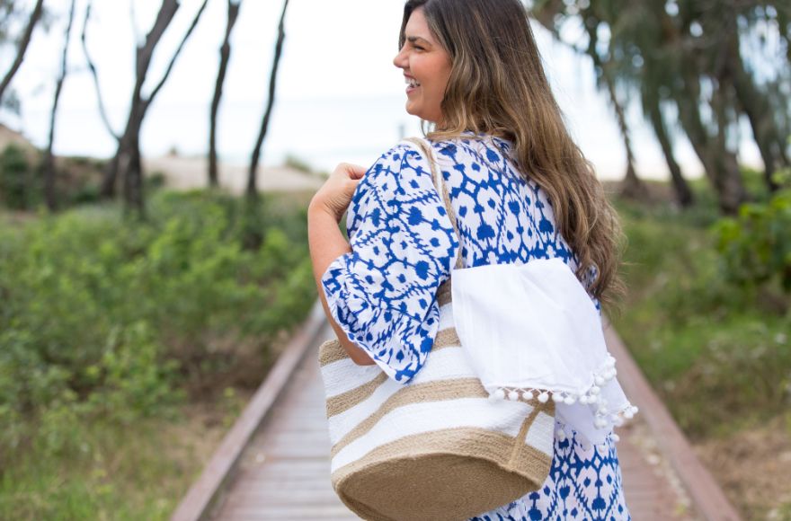 Woman with long brown hair wears blue and white kaftan and holds a jute and white striped beach bag with a white sarong inside it.