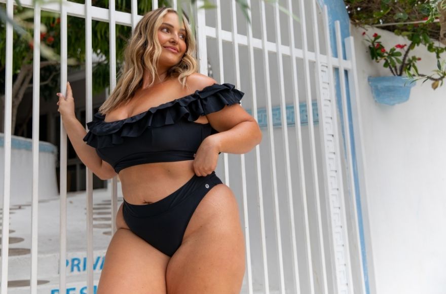Blonde woman poses by a white fence wearing a black frill off the shoulder bikini top and high waisted cheeky cut pant
