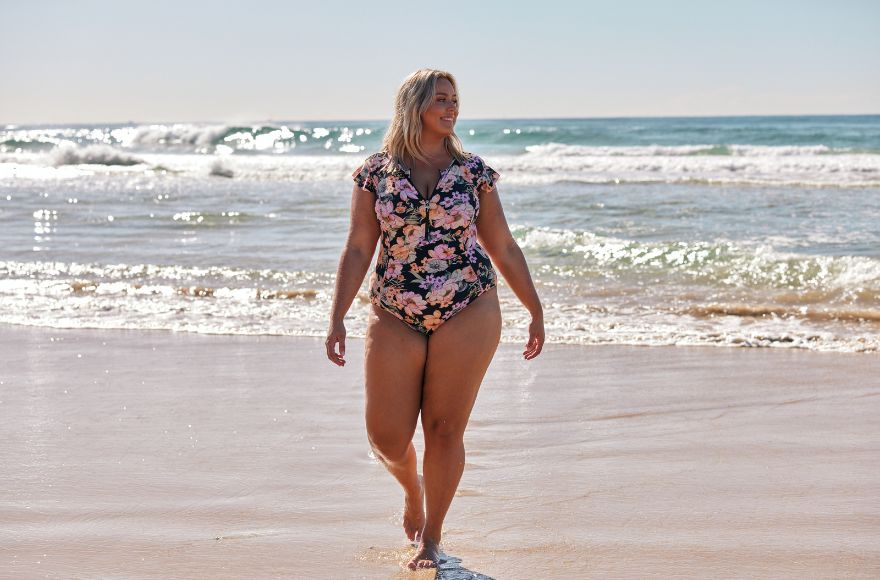 Woman with blonde hair walks along the beach wearing black and pink frill sleeve one piece swimsuit