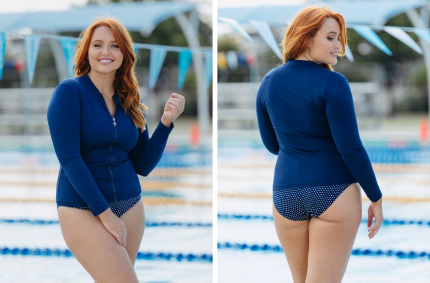 Woman with red hair poses by the pool wearing a long sleeve navy rash vest with a navy and white polkadot one piece