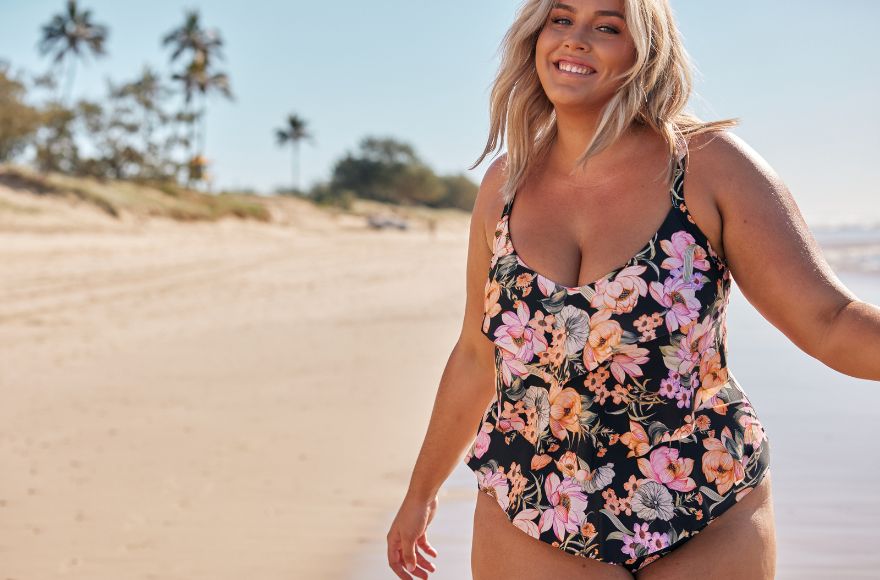 Woman with blonde hair poses on the beach wearing a frill one piece swimsuit. It has a black base with pink and orange floral print.