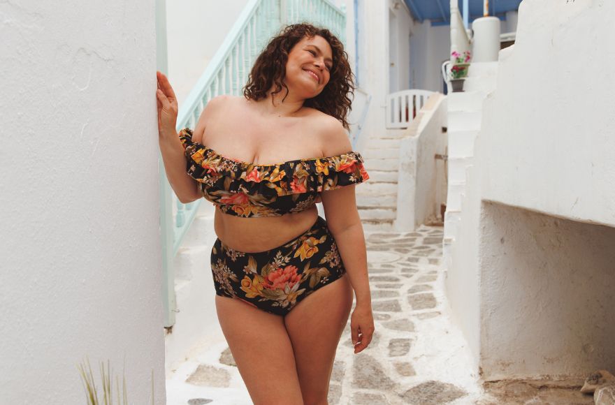 Brunette woman poses by a blue staircase wearing Frenchy Black floral double frill off the shoulder bikini top and high waisted bikini bottoms