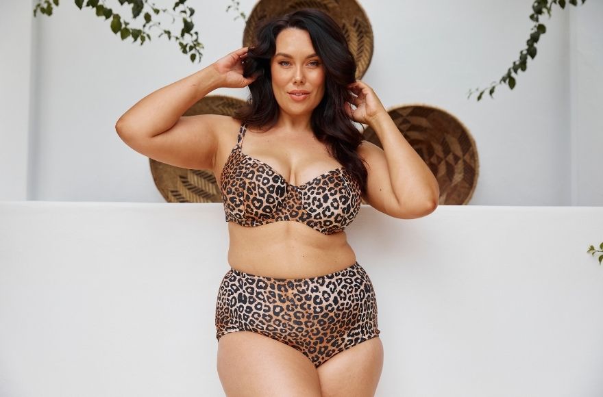Brunette woman poses against a white wall wearing Leopard print underwire bikini top and high waisted pant. 