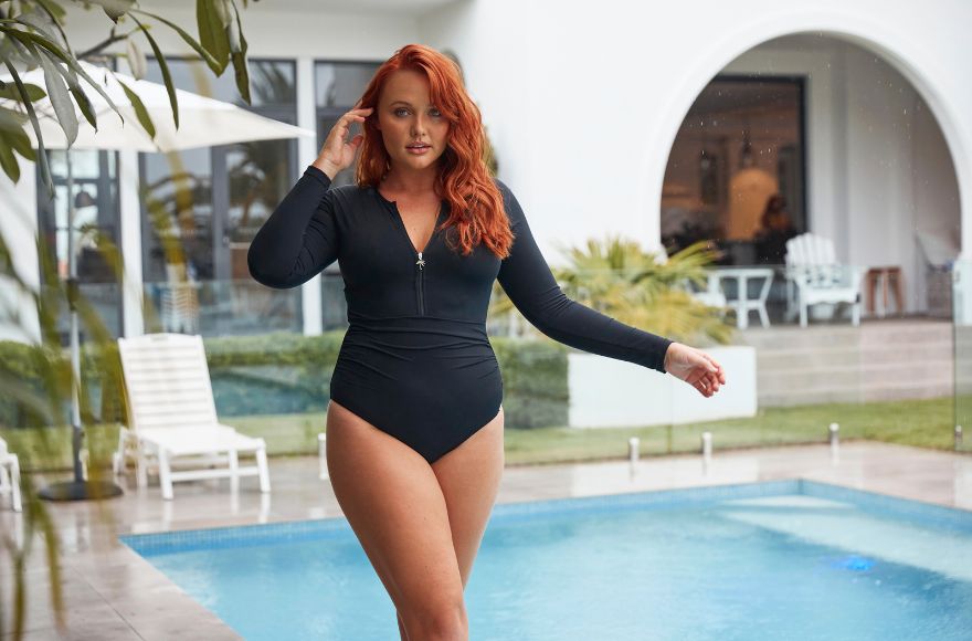 Woman with red hair poses by the pool wearing a black long sleeve one piece swimsuit