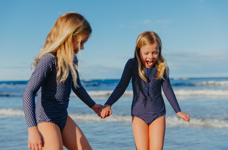 Two girls with long blonde hair play at the beach wearing navy and white dots long sleeve zip up one piece