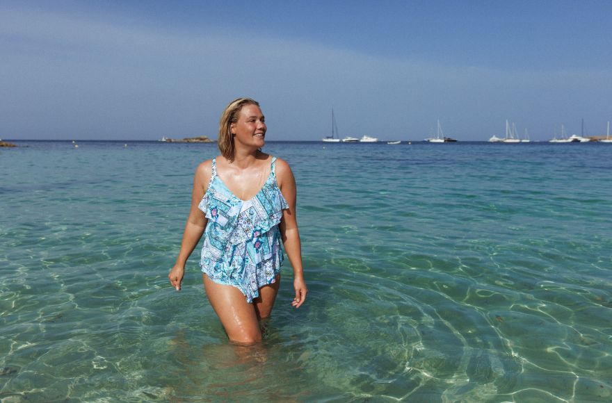 Woman with short blonde hair wades through the water wearing blue patchwork print ruffled tankini top