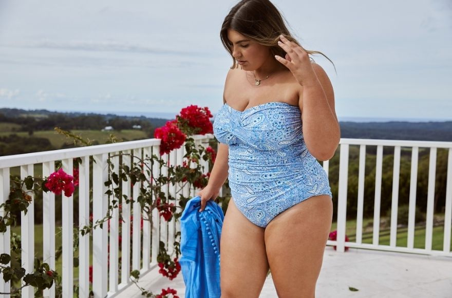Brunette model poses on a balcony wearing Santorini blue paisley twist front bandeau strapless one piece swimsuit
