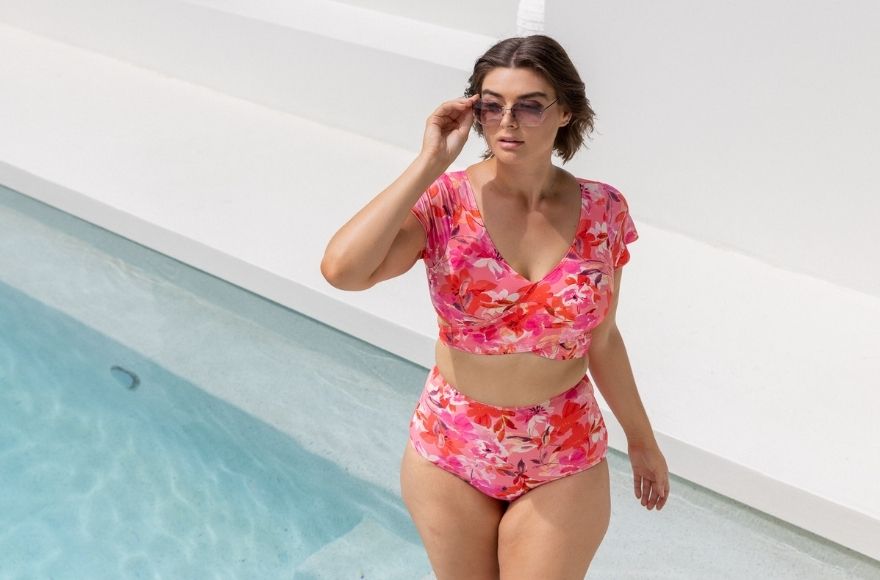 Brunette model poses by the pool wearing Pink Bloom crop wrap top and high waisted pants