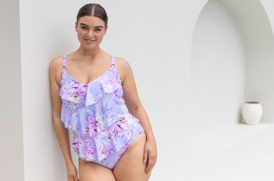 brunette woman leans against white wall wearing Lilac Florence 3 tier ruffle one piece swimsuit