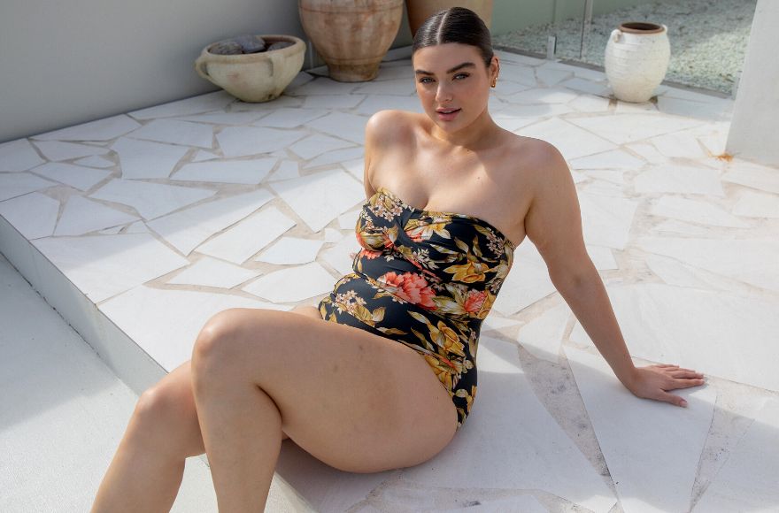 Woman with slicked back brown hair lounges by the pool wearing Frenchy Black floral strapless twist front bandeau one piece swimsuit.