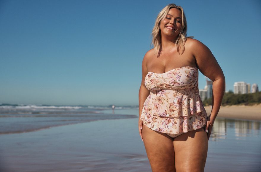 Woman with blonde hair poses on the beach wearing a strapless frilled tankini top in a pale pink floral print