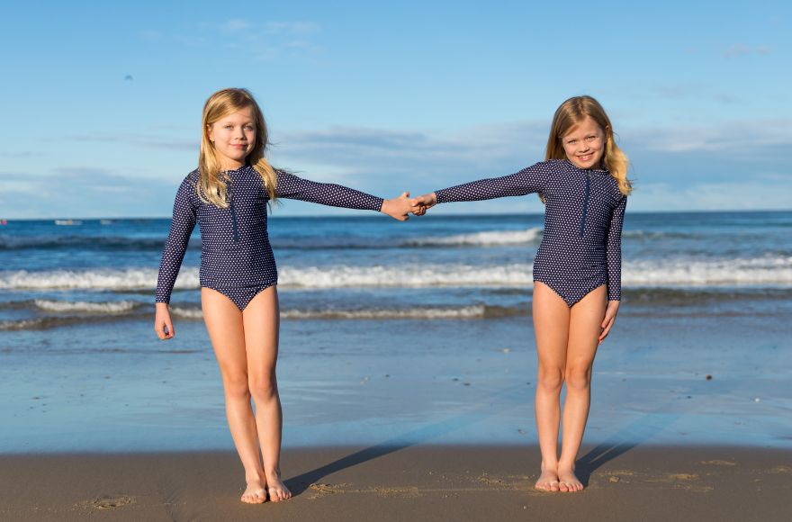 Girls with blonde hair hold hands at the beach wearing long sleeve navy one piece swimsuits with white polka dots.