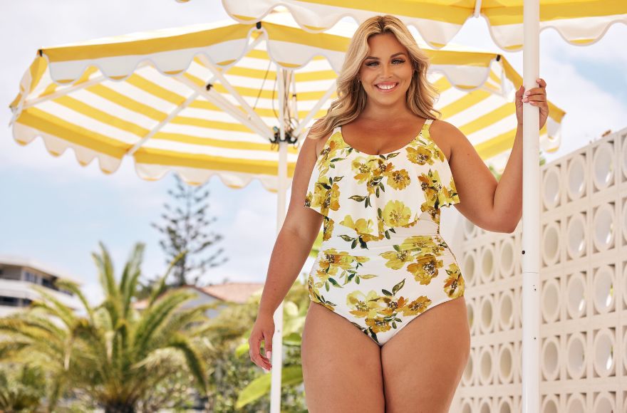 Woman with blonde hair stands under a yellow and white striped umbrella. She wears a white one piece swimsuit with a yellow floral print.