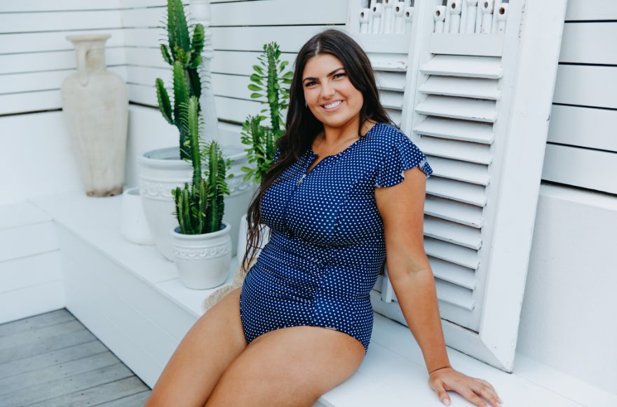 Woman with long dark brown hair sits on a white bench wearing a navy one piece with white polkadots.