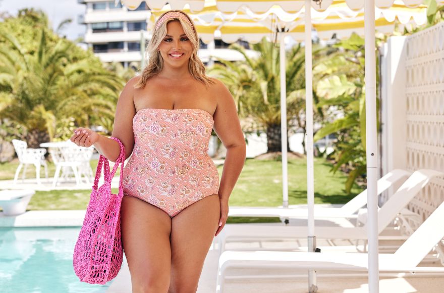 Woman with blonde hair poses by the pool and yellow and white stripe umbrellas wearing a pink floral strapless one piece. She is carrying a bright pink woven straw bag