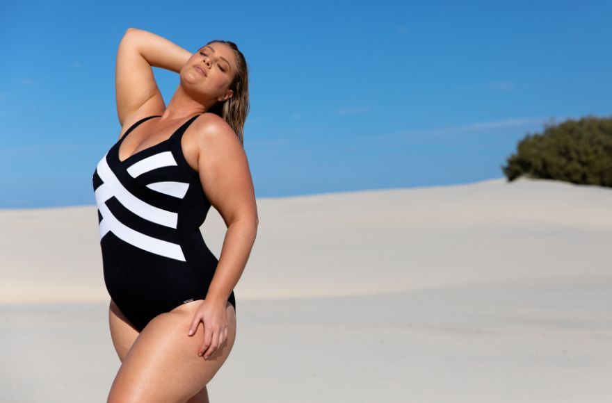 Woman with blonde hair poses on the beach wearing black and white criss cross one piece swimsuit