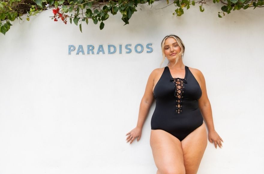 blonde woman leaning against a white wall wearing a black lace up one piece swimsuit