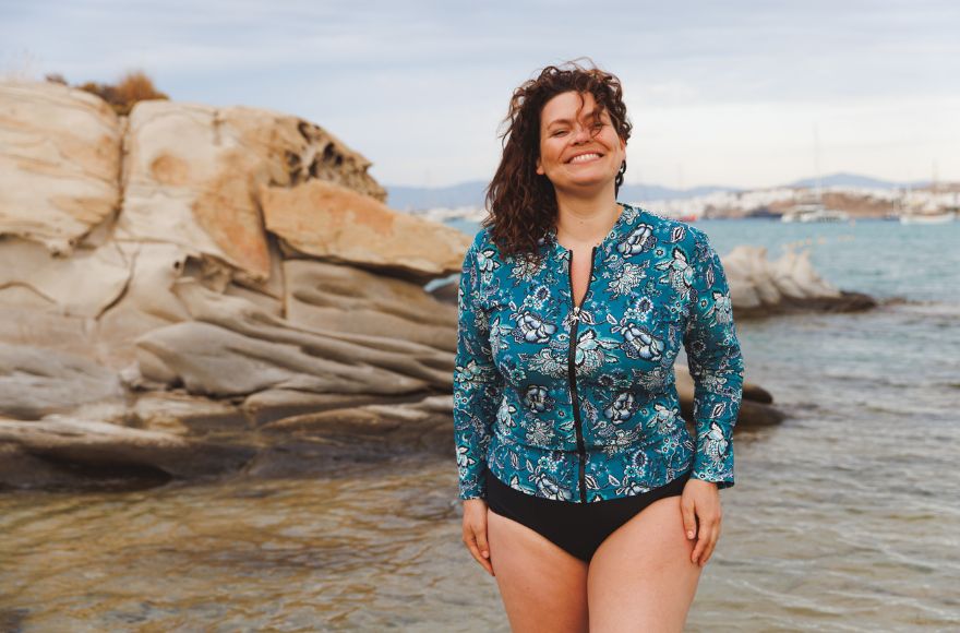 Woman with brown hair poses at the beach wearing Sardinia teal floral long sleeve zip up rash vest and black pants