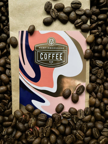 Image of a bag of Martinborough Coffee with a pink, purple, and white pattern on the label. The bag of coffee is surrounded by roasted coffee beans.