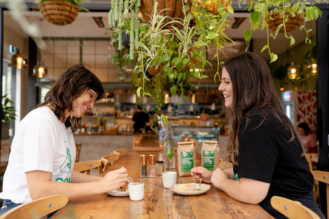 Two friends are facing each other at a long table in a cafe, smiling and talking over some delicious common good coffee
