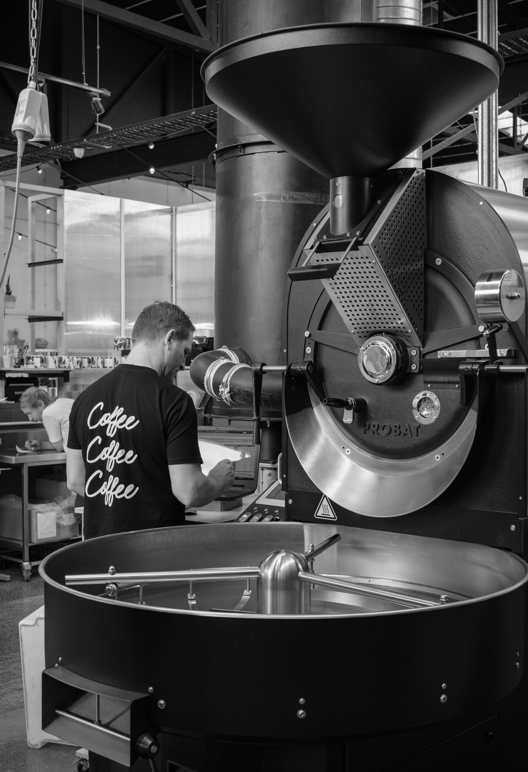 Black & white image of head roaster Andy standing next to a large Probat P25 roasting drum, his back to the camera.
