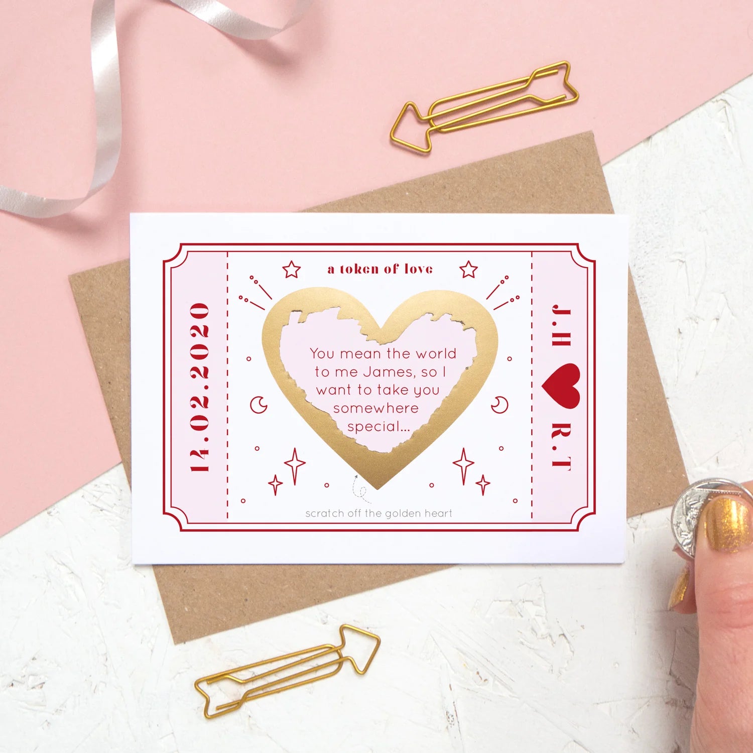 A love token scratch card which has been personalised with a special message, date and initials. It has been photographed on a pink and white background and the gold heart on the card has been scratched off to reveal the message.