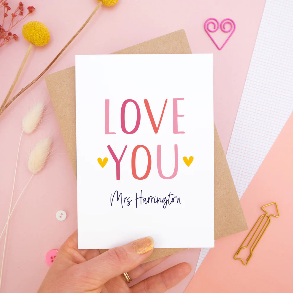 A personalised 'love you' card with the recipients name. This card has been shot on a pink background surrounded by stationery and dry flowers.