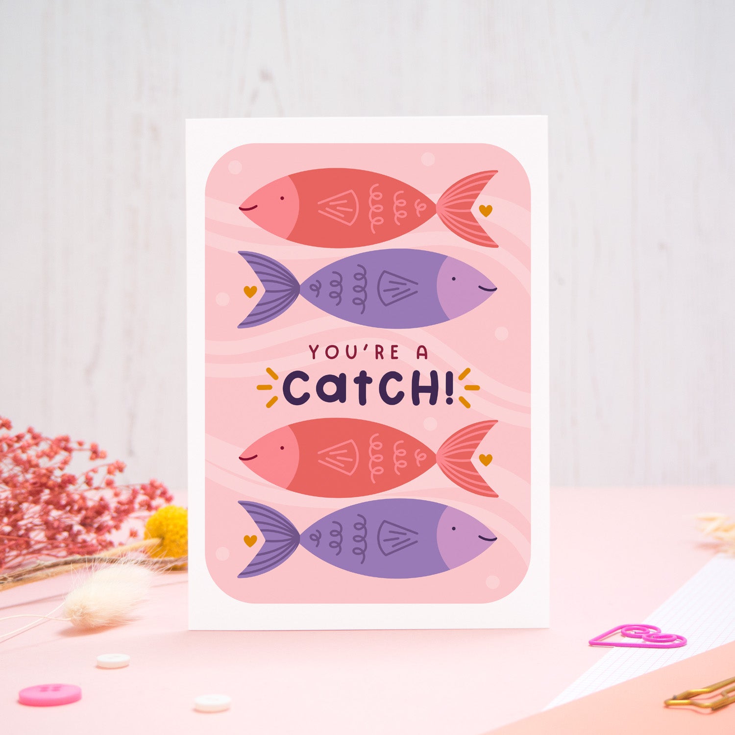 You're a catch valentine's day card featuring pink and purple fish. The card has been photographed on a pink and white background with stationery and dry flowers surrounding it.