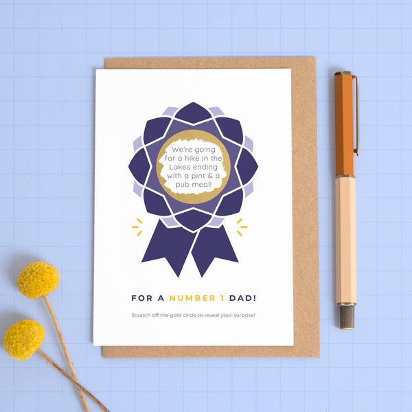 A scratch card for dad designed as a rosette! Scratch out the centre to reveal the surprise! This is the navy and purple version photographed on a blue background.