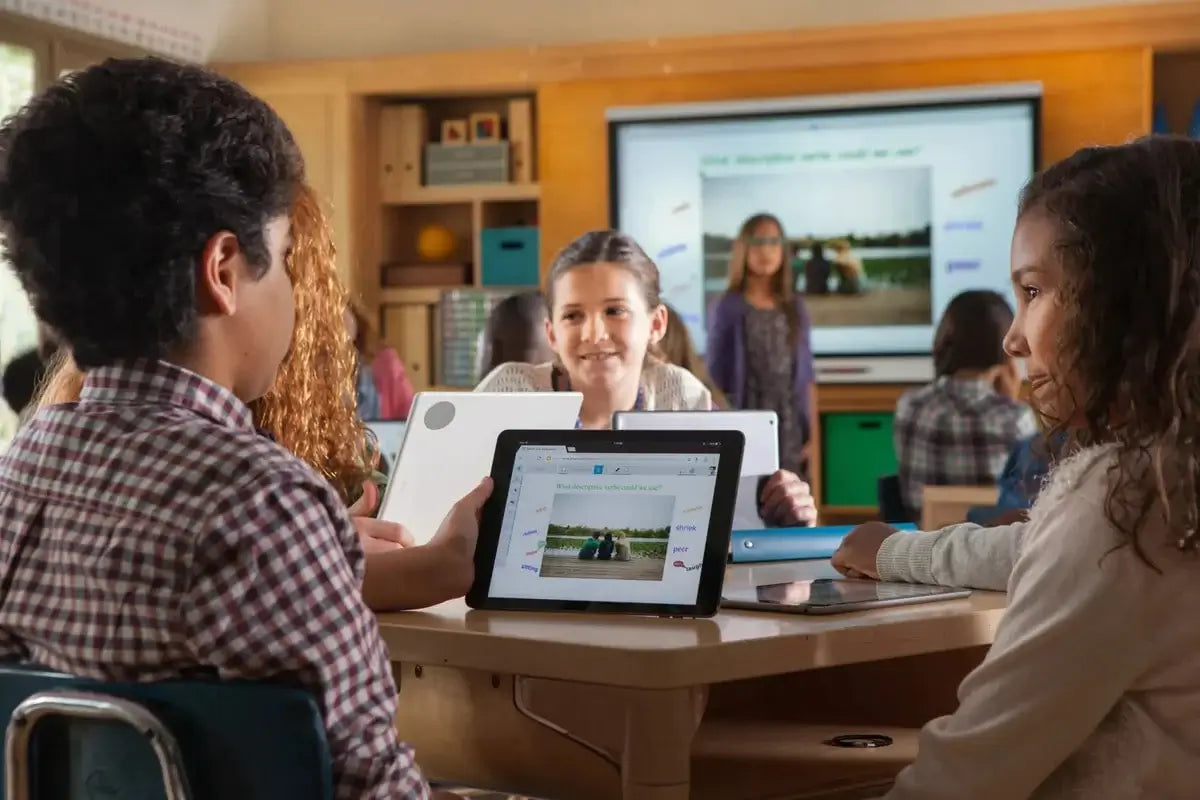 Three young children doing school work on tablets with a teacher working on a SMART Board interactive panel in the background 