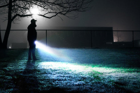 guy with a flashlight walking a grassy field at night near a chainlink fence
