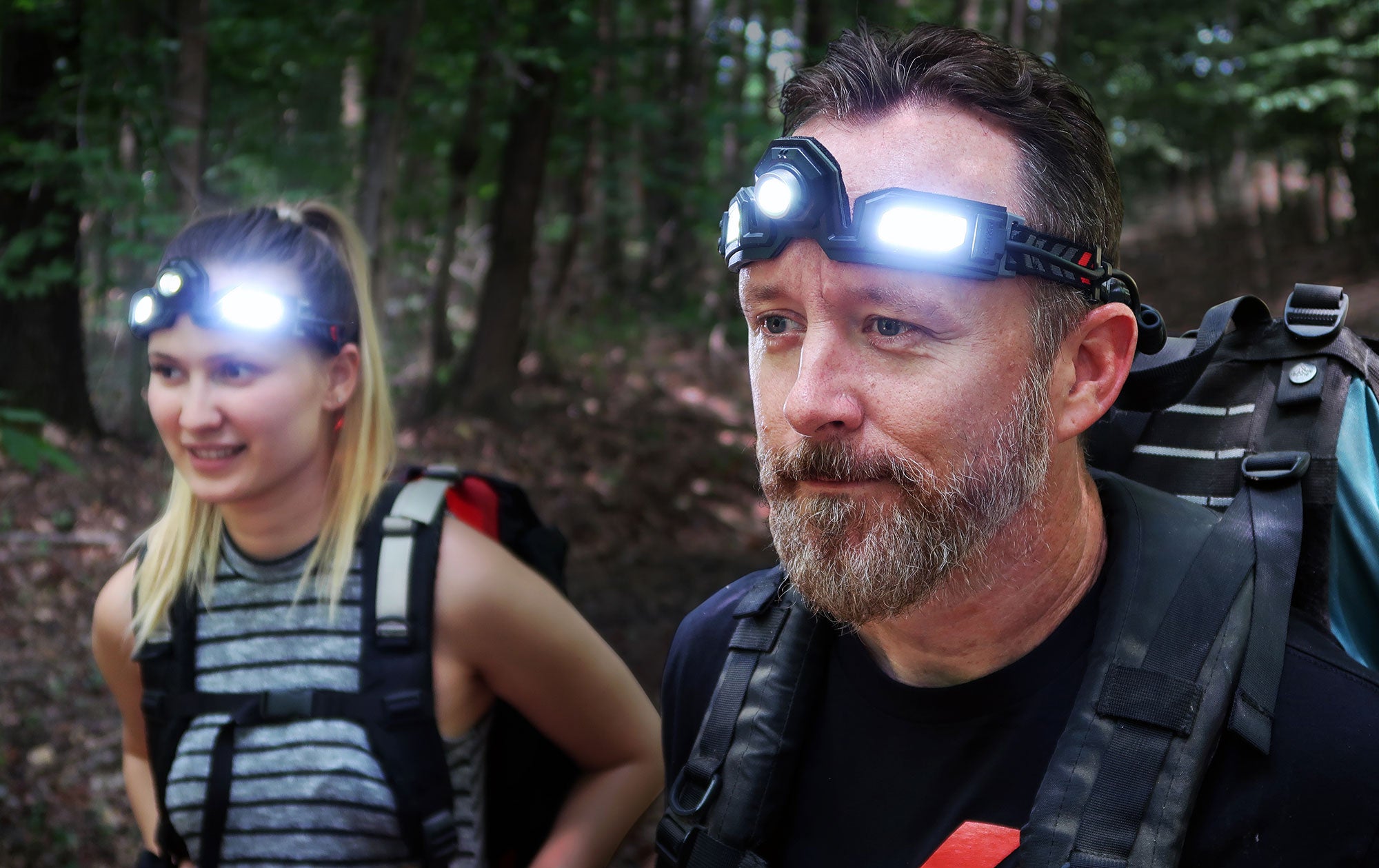 Close up lifestyle image of a man and woman out in the woods backpacking/hiking with headlamp pros on.