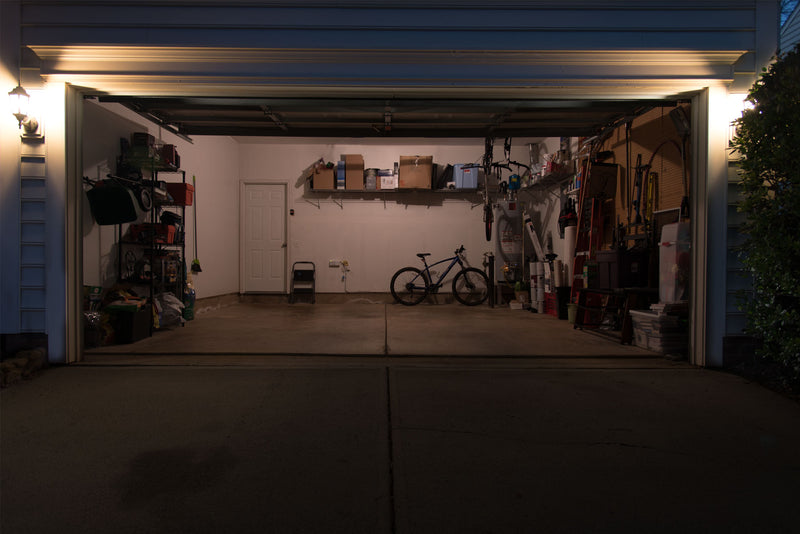 2 car garage with lots of stuff stored inside around the outside of the room. Currently lit by a single incandescent standard light bulb.