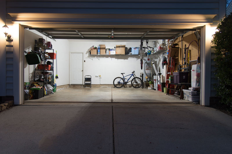 2 car garage with lots of stuff stored inside around the outside of the room. Currently lit by a single TRiLIGHT 3000 lumen deformable  light bulb.